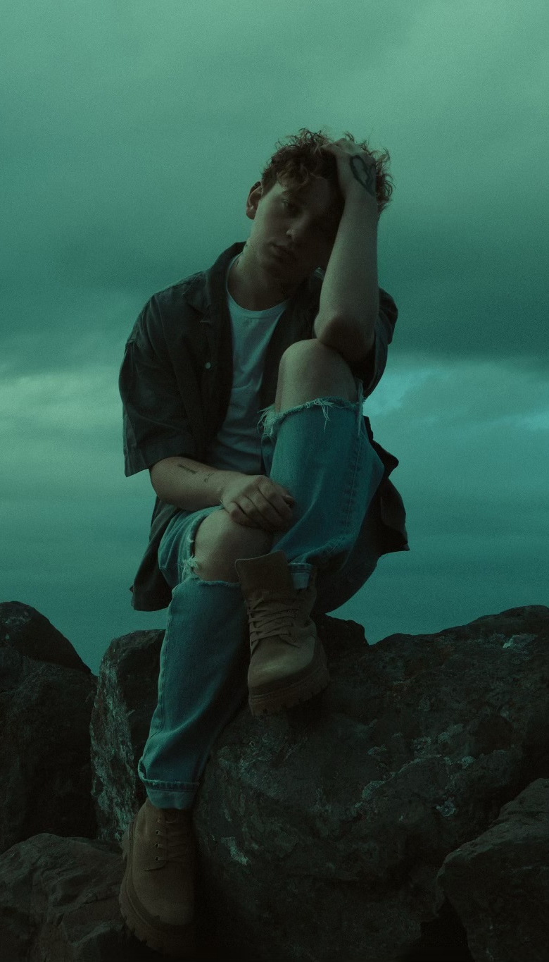 Shaya Zamora sits on large rocks with a hand resting on his head. The background features a cloudy, green-tinted sky, giving the image a moody atmosphere.